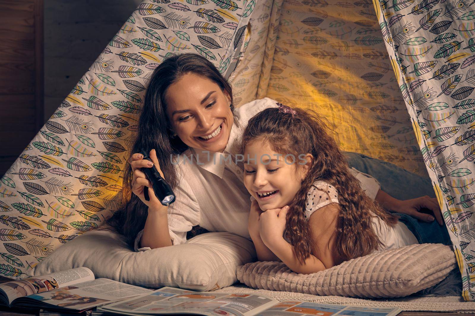 Mother and daughter are sitting in a teepee tent, reading stories with the flashlight. Happy family. by nazarovsergey