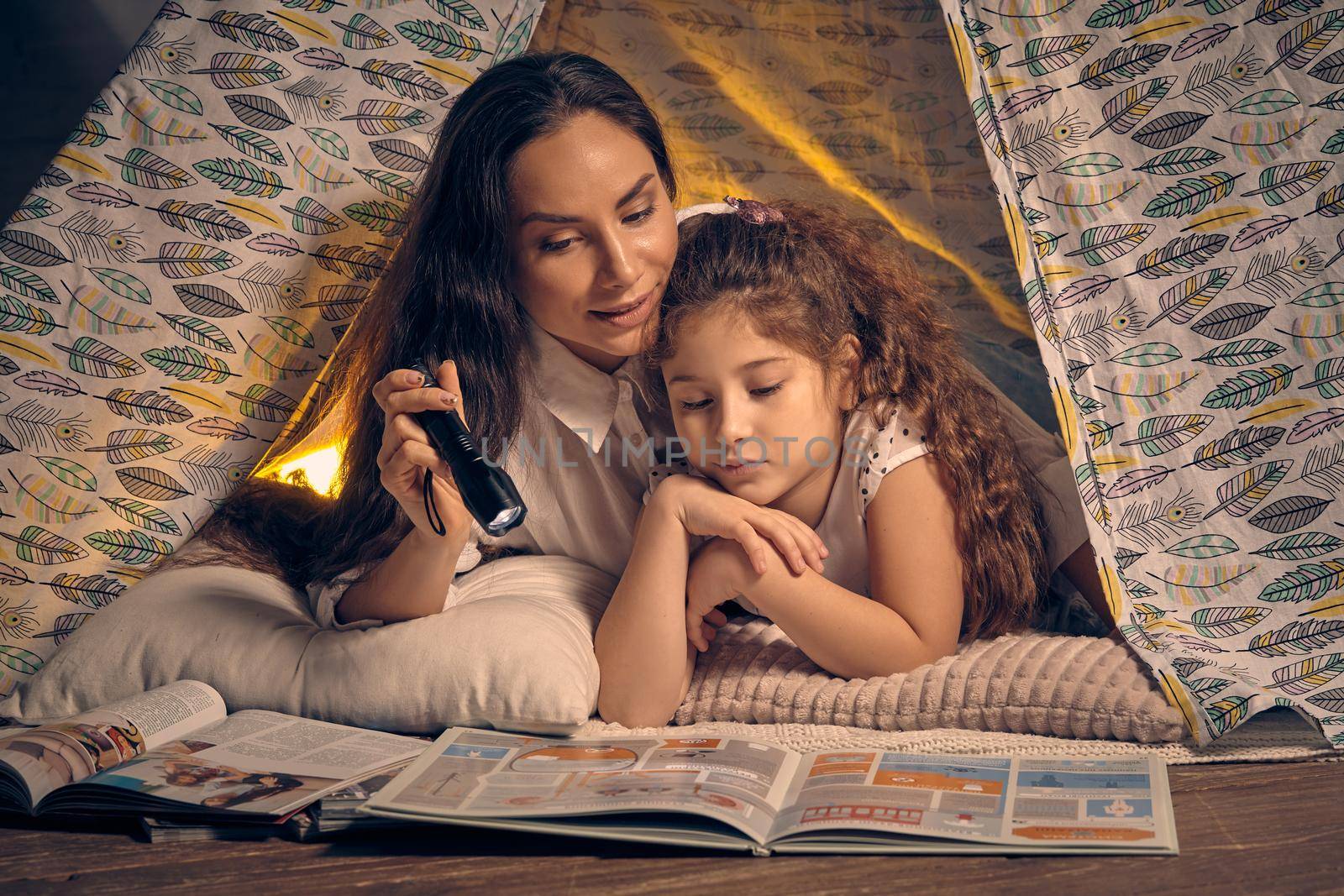 Mother and her charming daughter are sitting in a teepee tent, reading stories with the flashlight. Happy family.