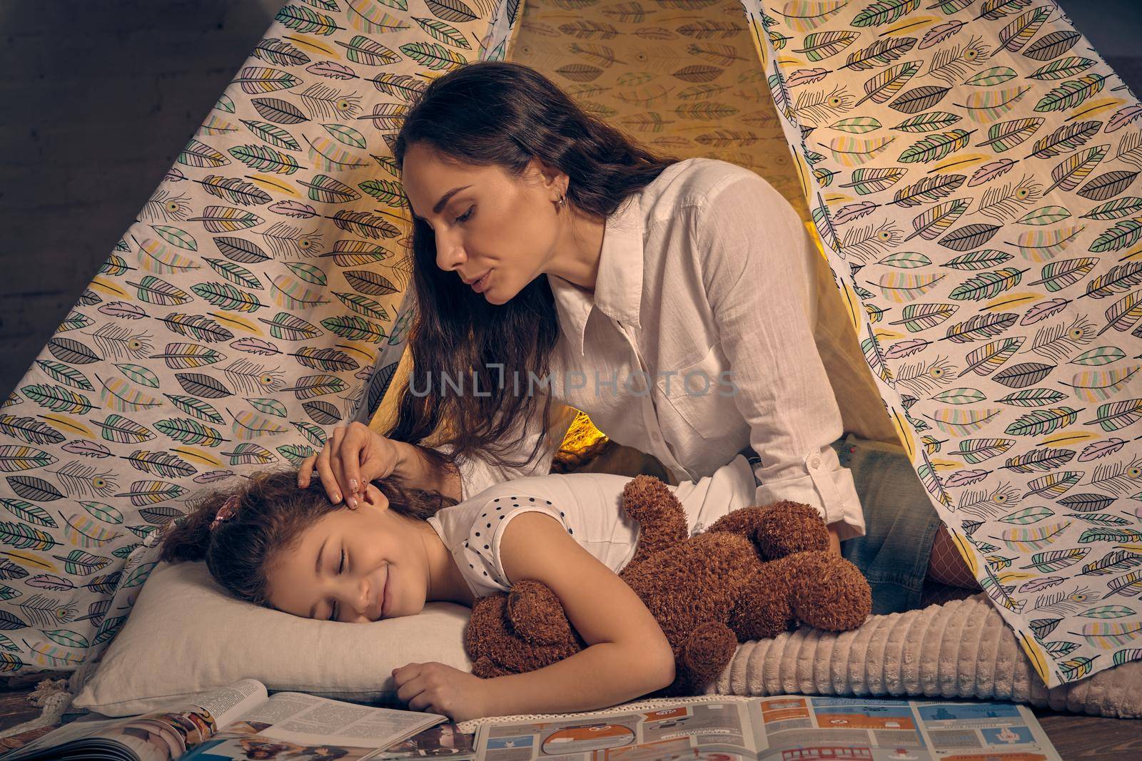 Beautiful mother and her pretty daughter are in a teepee tent with some pillows. Girl is sleeping and hugging a toy bear. Mom is looking and stroking her. Happy family.