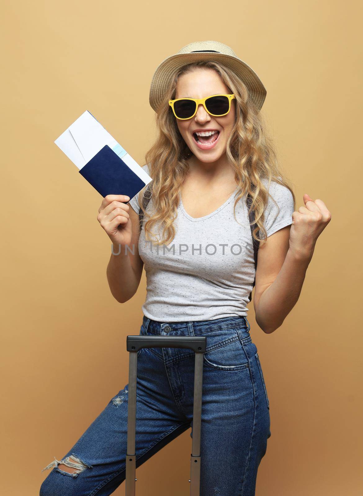 Cheerful blonde woman in sunglasses posing with baggage and holding passport with tickets over beige background. by tsyhun