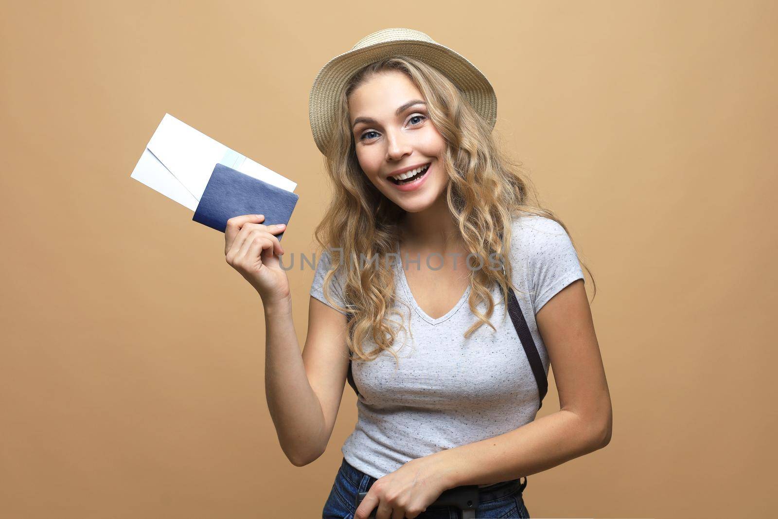 Beautiful blonde woman wearing summer clothes posing with passport with tickets over beige background