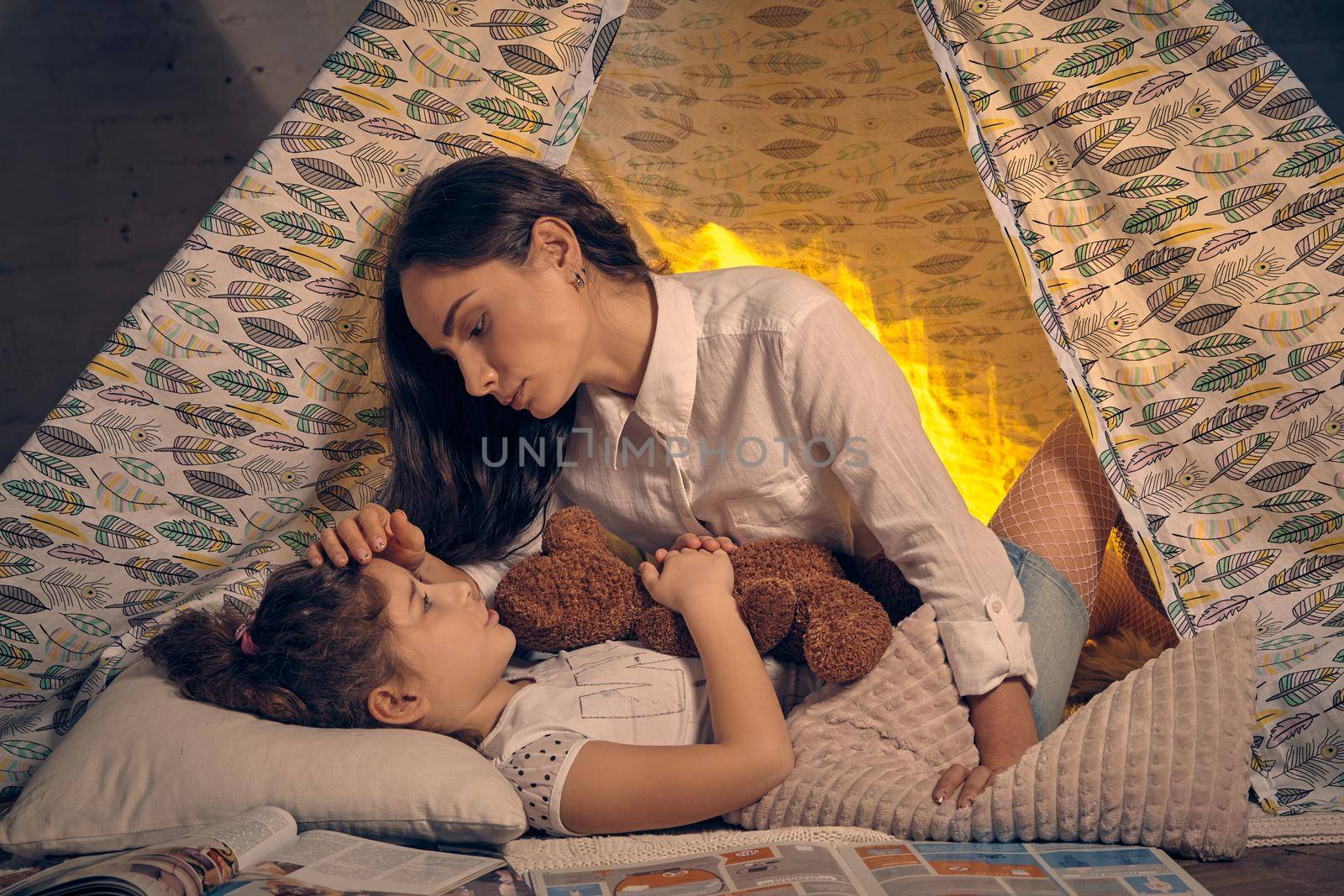 Mother and her daughter are in a teepee tent with some pillows. Girl is lying and hugging a toy bear. They are looking at each other and mom is stroking her. Happy family.