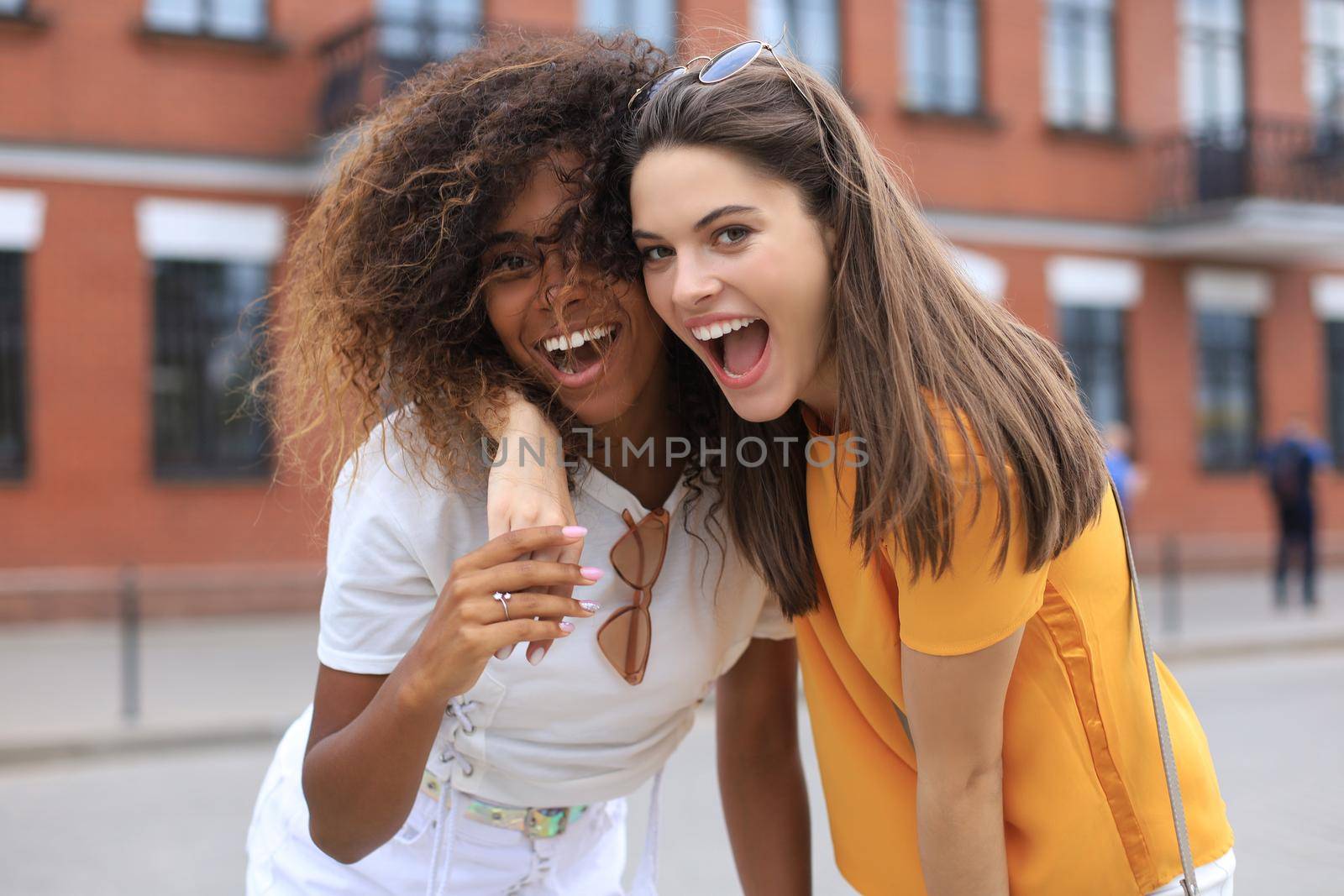 Two young smiling hipster women in summer clothes posing on street.Female showing positive face emotions