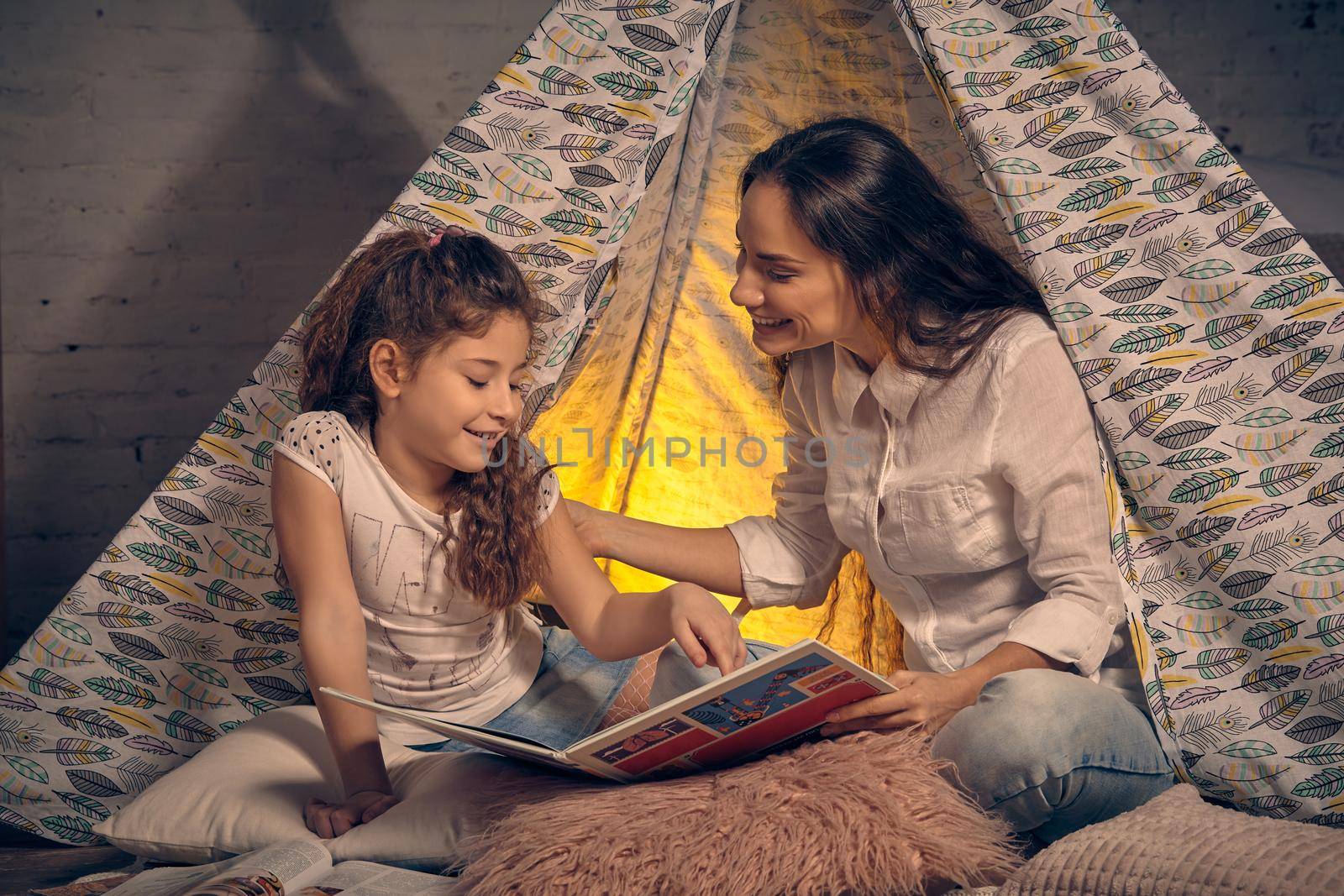 Mother and daughter are sitting in a teepee tent with some pillows, reading stories and smiling. Happy family.