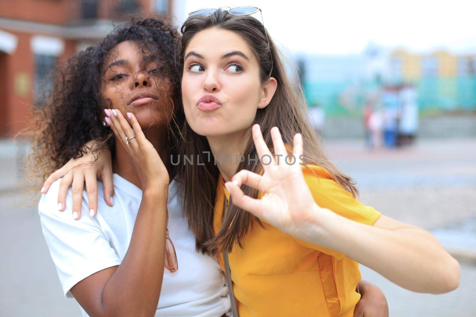 Two young smiling hipster women in summer clothes posing on street.Female showing positive face emotions