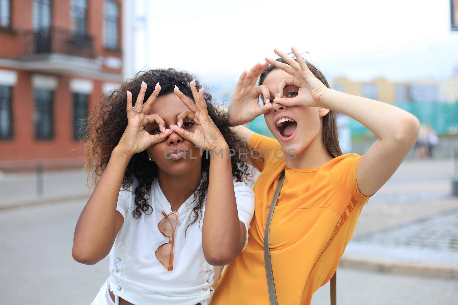 Two young smiling hipster women in summer clothes posing on street.Female showing positive face emotions