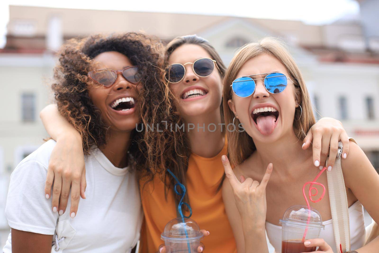 Three young smiling hipster women in summer clothes posing on street.Female showing positive face emotions