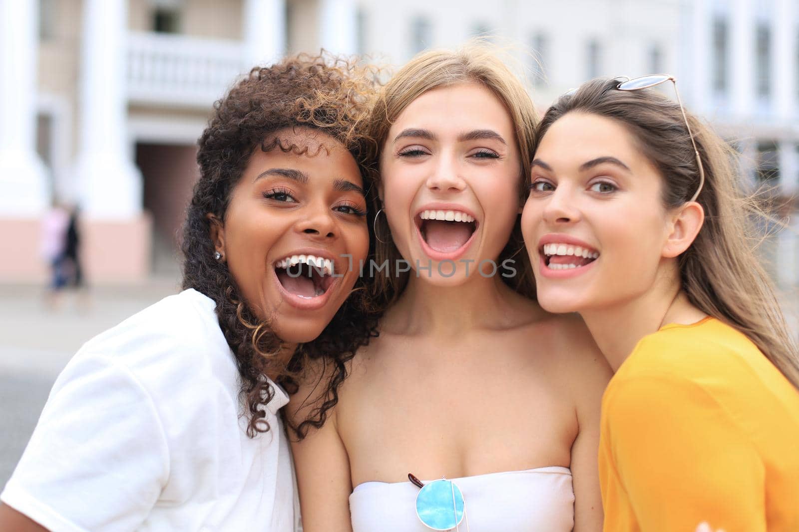 Three young smiling hipster women in summer clothes posing on street.Female showing positive face emotions