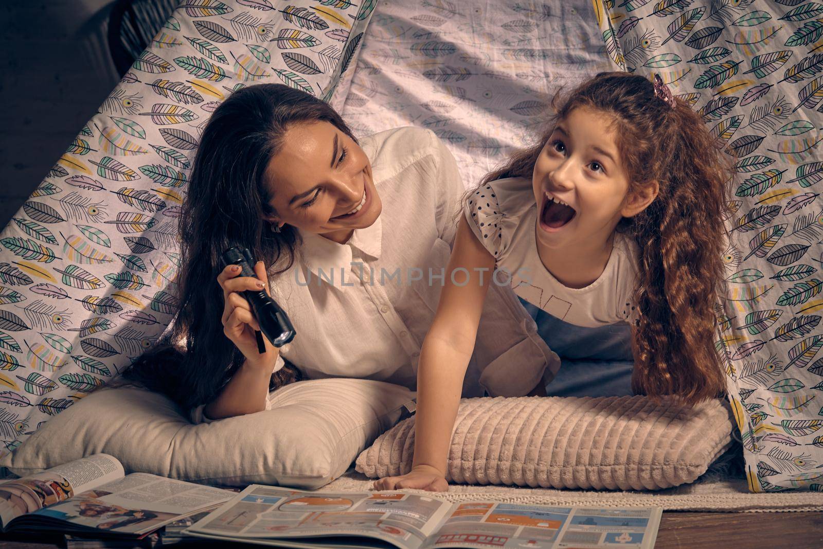 Mother and daughter are sitting in a teepee tent, reading stories with the flashlight. Happy family. by nazarovsergey