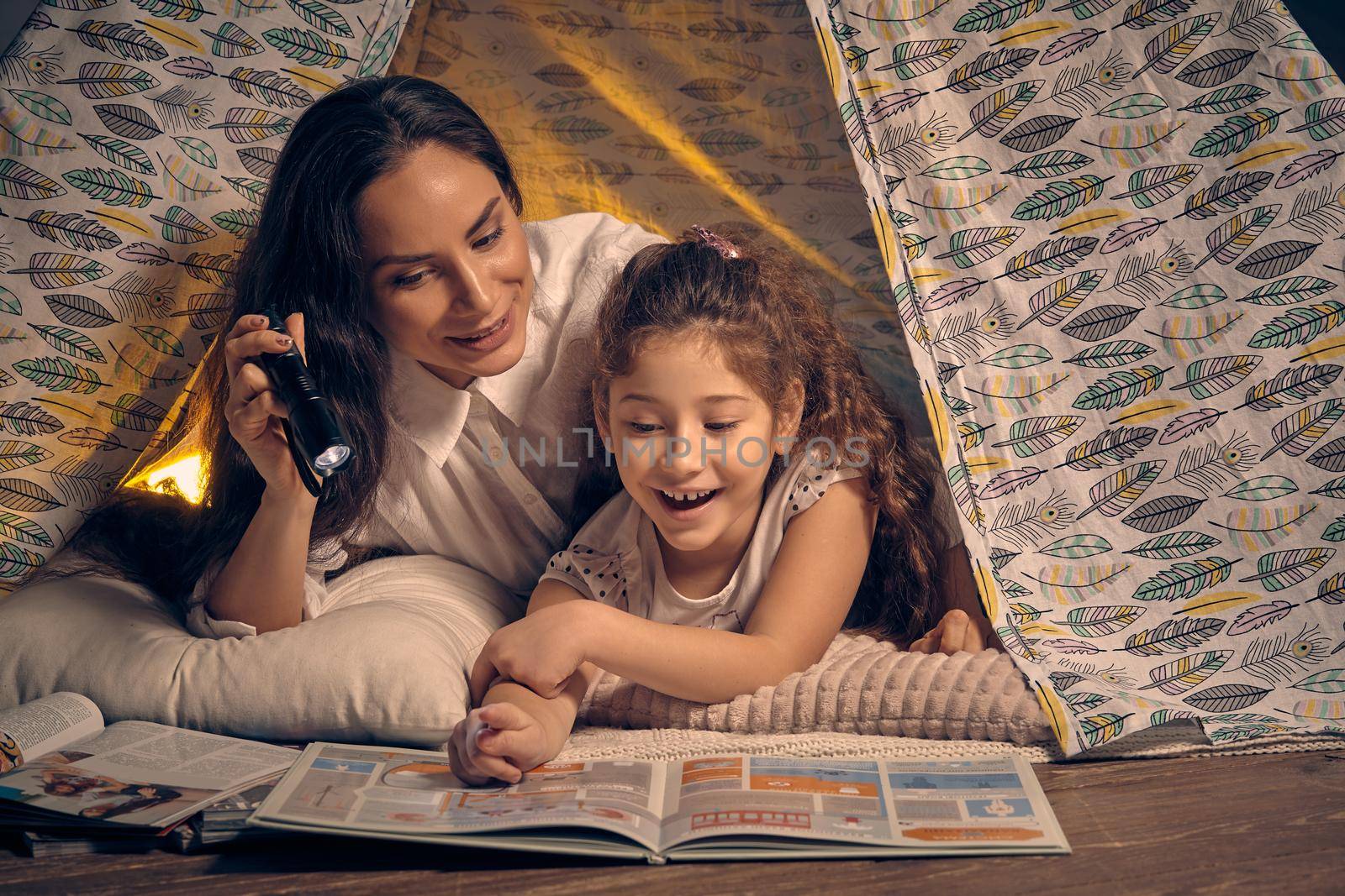 Mother and her charming daughter are sitting in a teepee tent, reading stories with the flashlight and smiling. Happy family.