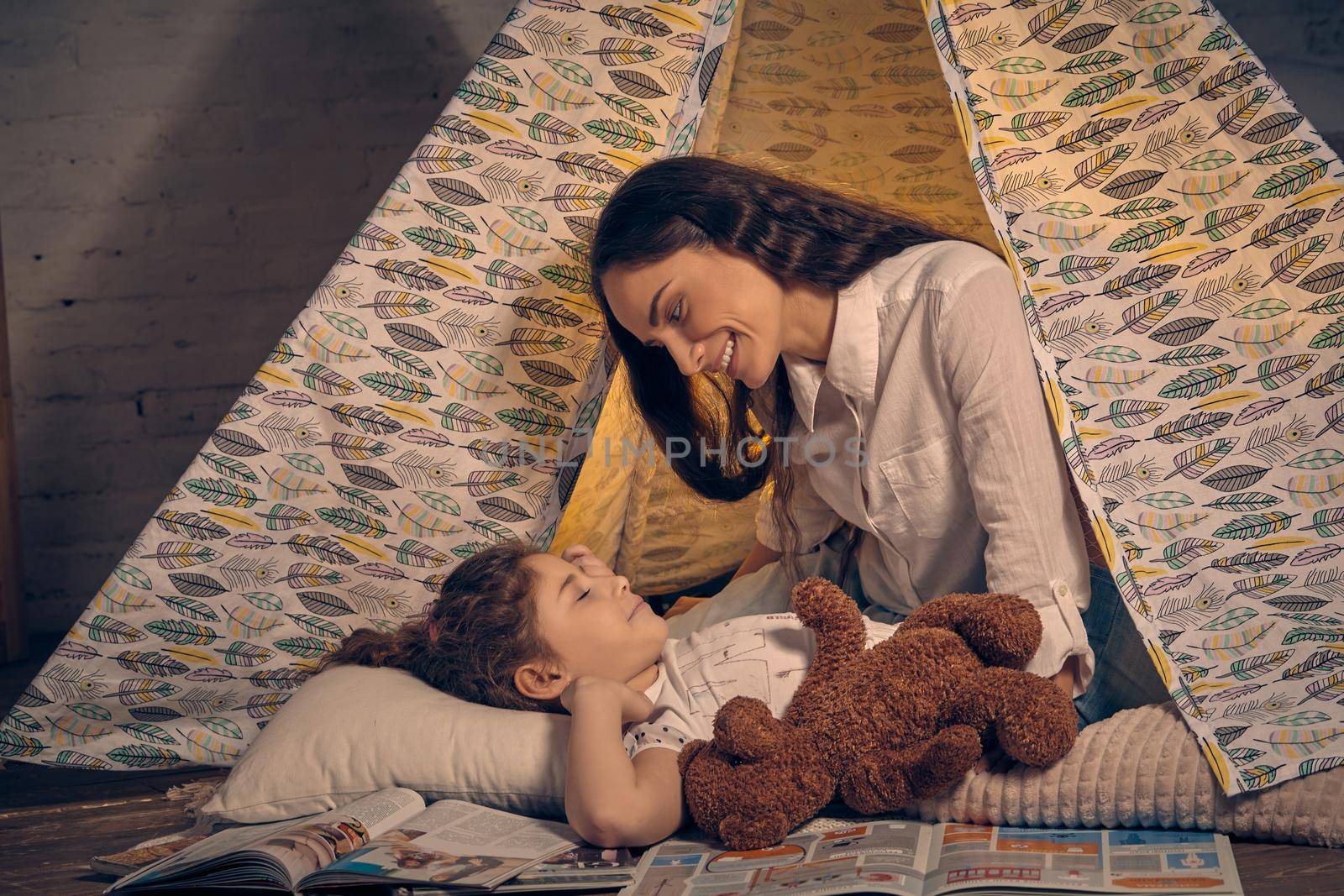 Young mother and her sweet daughter are in a teepee tent with some pillows. Girl is lying with a toy bear. Mom is looking at her. Happy family.