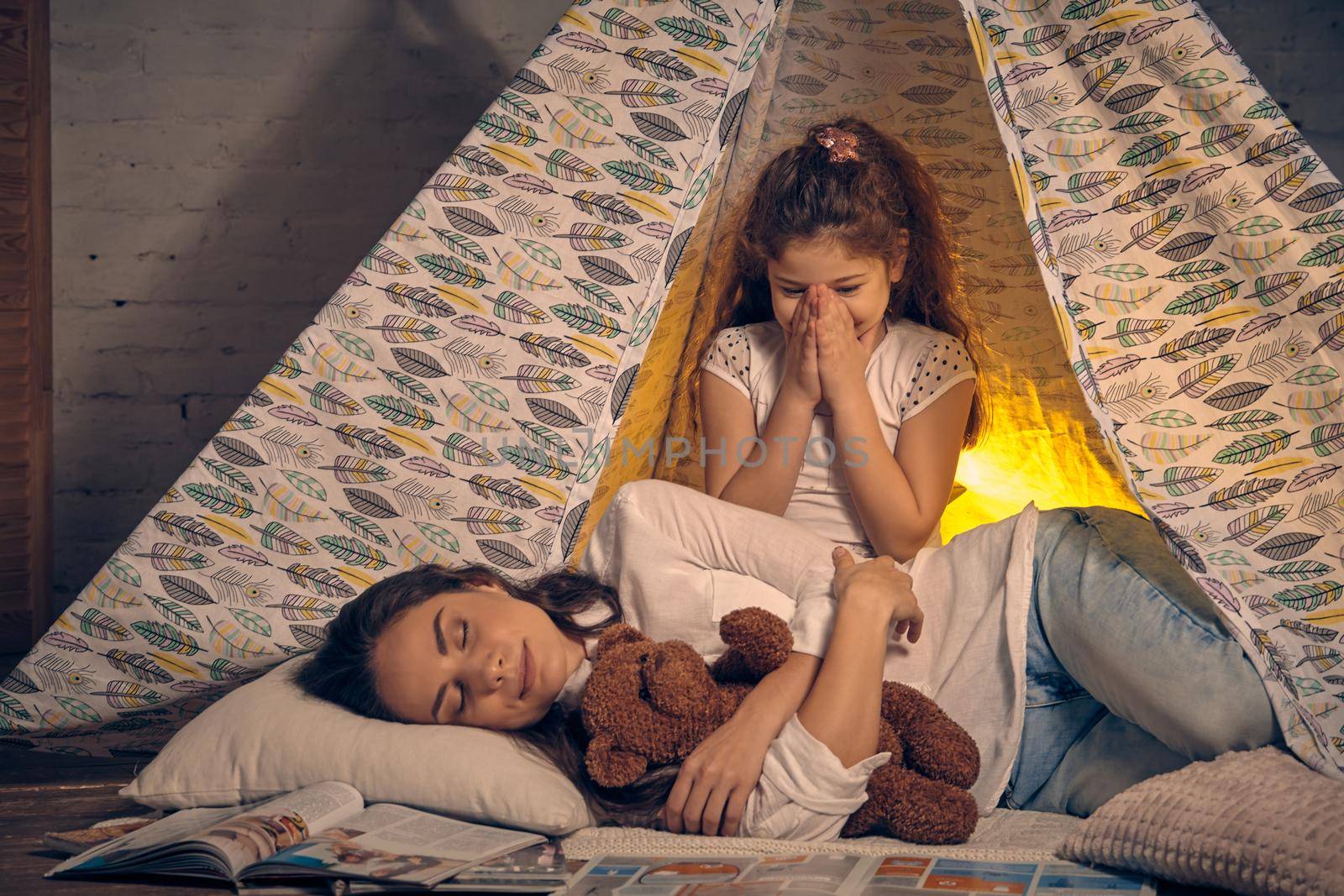 Young mother and her pretty daughter are in a teepee tent with some pillows. Mom is sleeping with a toy bear. Daughter is smiling and looking at her. Happy family.