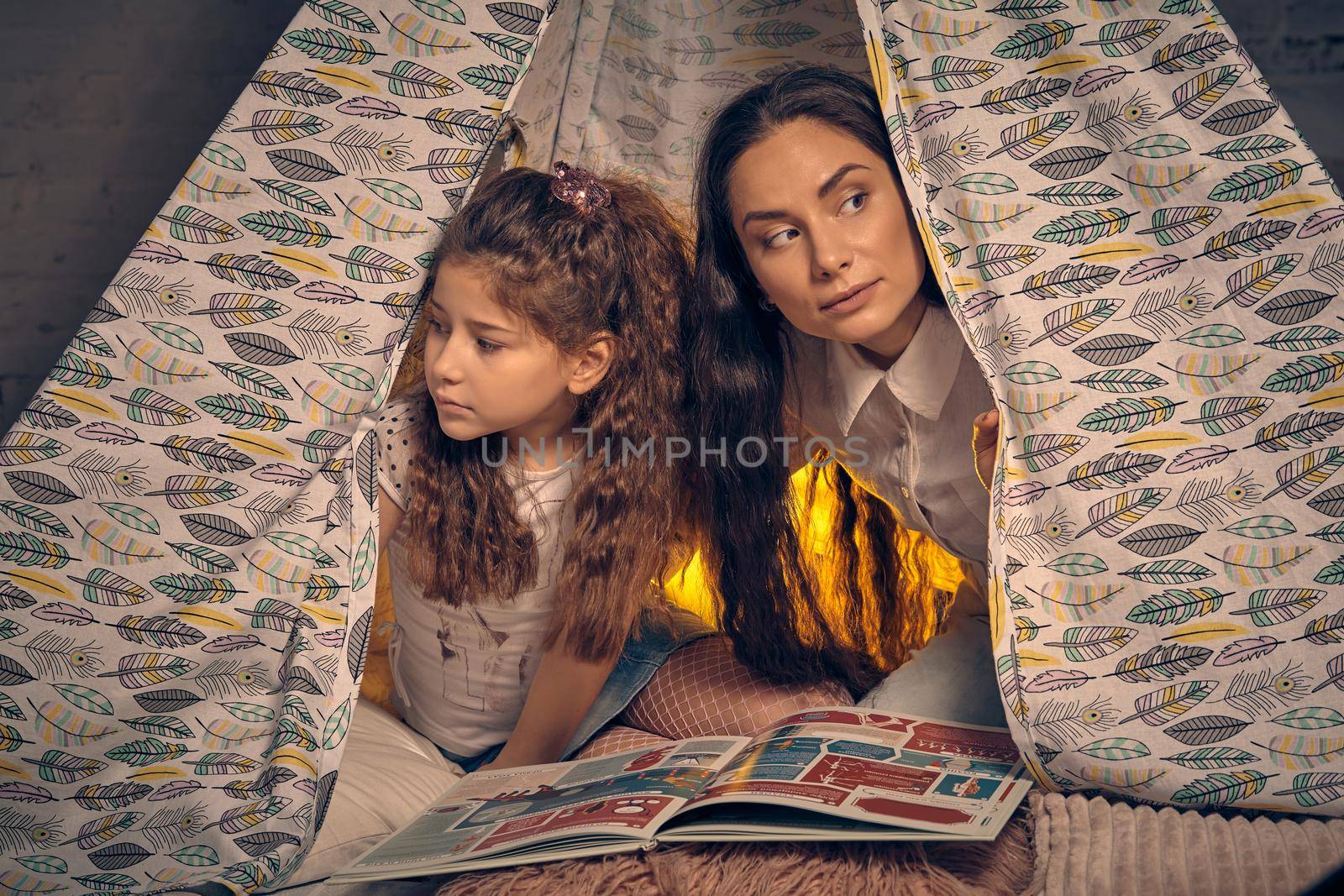 Young mother and her daughter are sitting in a teepee tent with some pillows and looking around. Happy family.