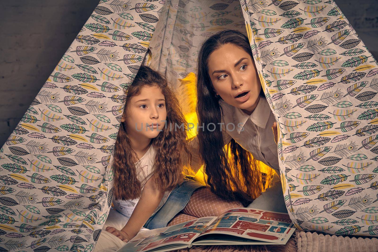 Mother and daughter are sitting in a teepee tent with some pillows and reading stories. Happy family. by nazarovsergey