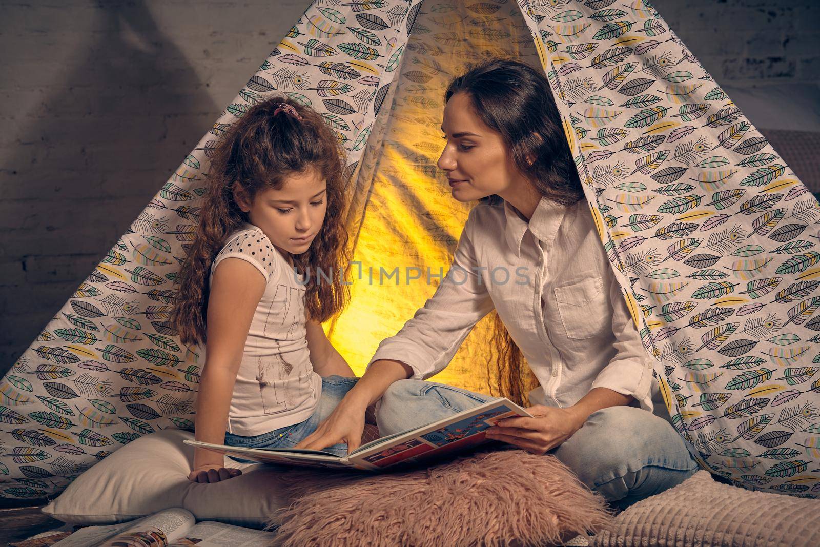 Mother and daughter are sitting in a teepee tent with some pillows and reading stories. Mom is showing something in the book and looking at her. Happy family.