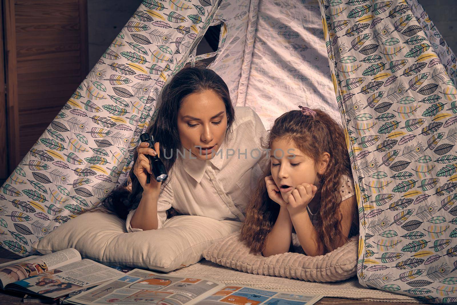 Mother and daughter are sitting in a teepee tent, reading stories with the flashlight. Happy family. by nazarovsergey