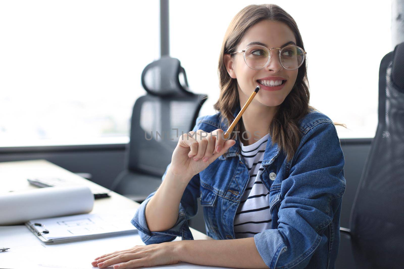 Beautiful young woman looking at camera and holding pencil while sitting in modern office