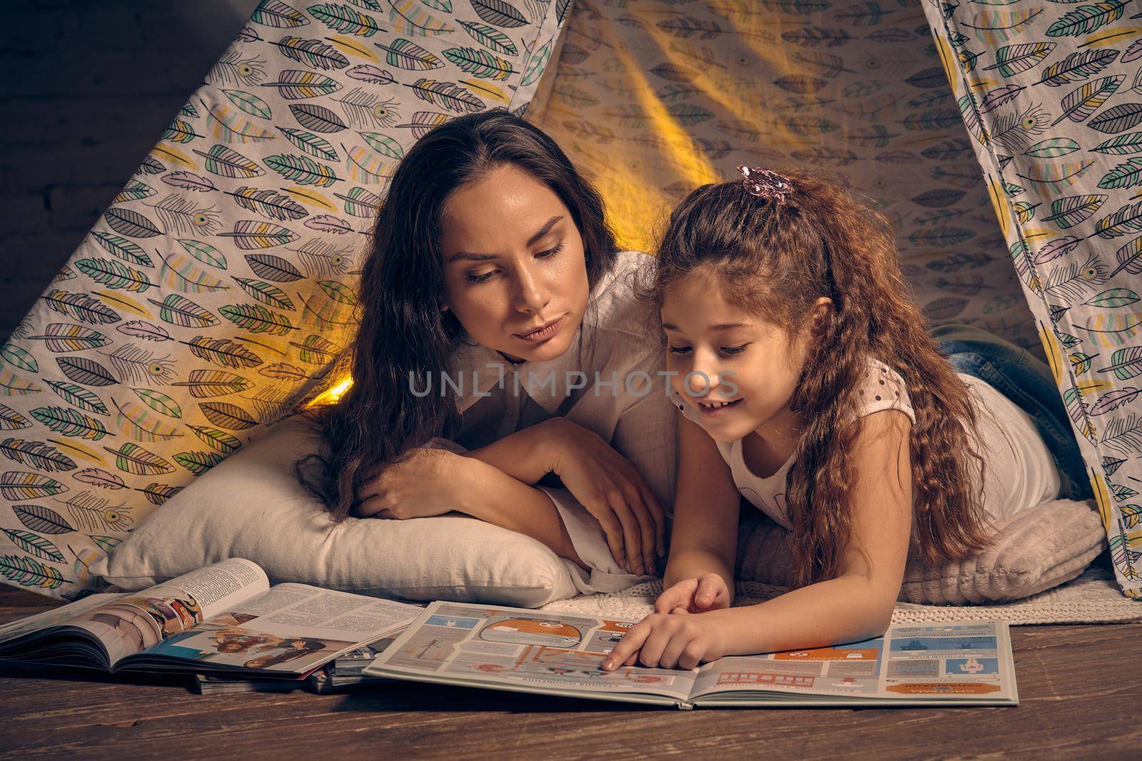 Mother and daughter are sitting in a teepee tent with some pillows and reading stories. Happy family. by nazarovsergey