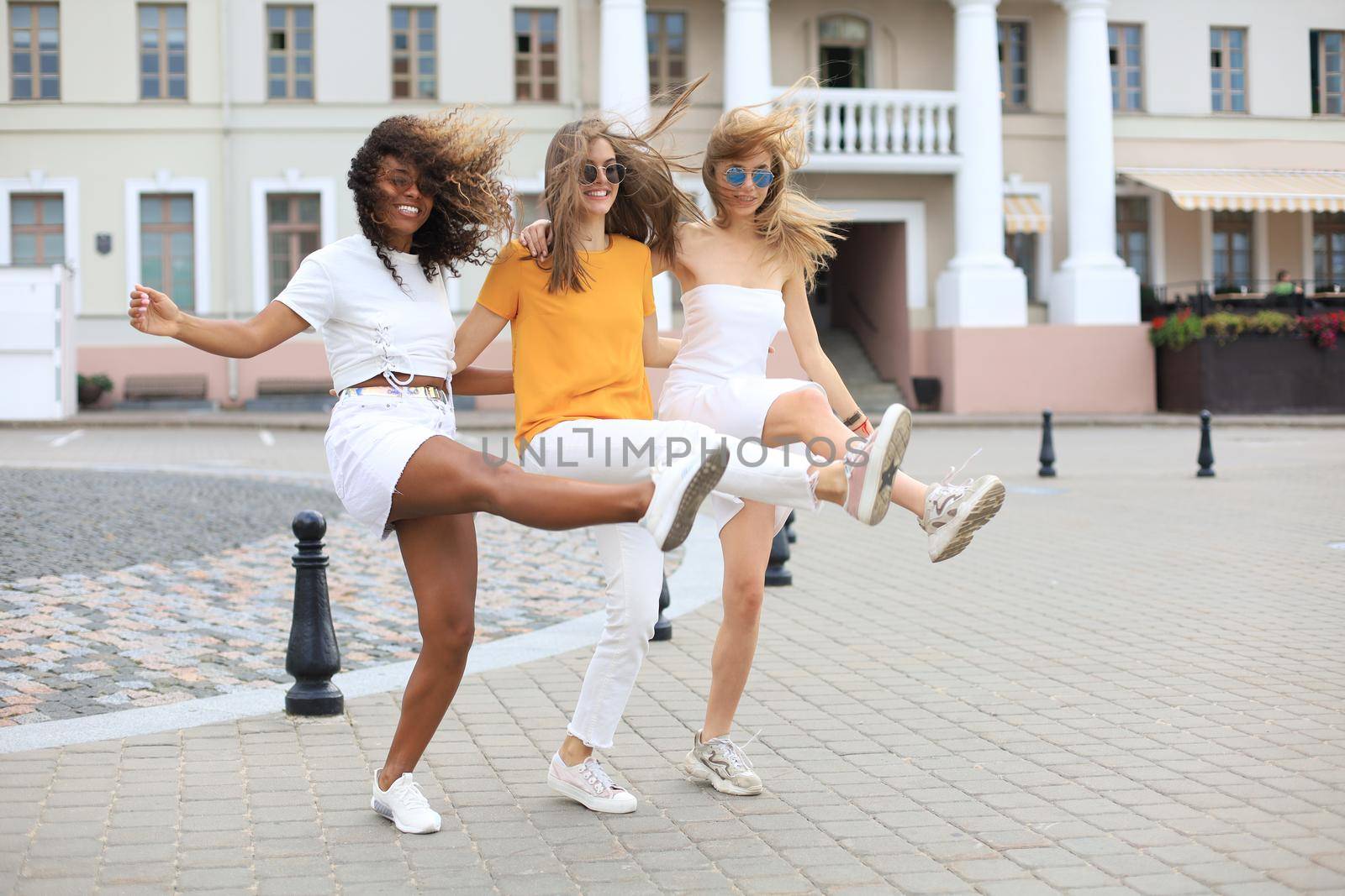 Three young smiling hipster women in summer clothes posing on street.Female showing positive face emotions. Dancing
