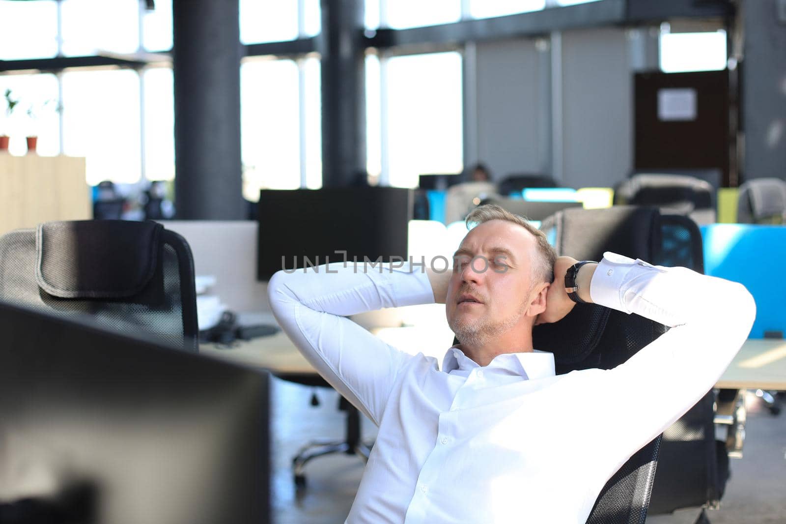 Middle aged modern businessman keeping hands behind head and smiling while sitting in the office