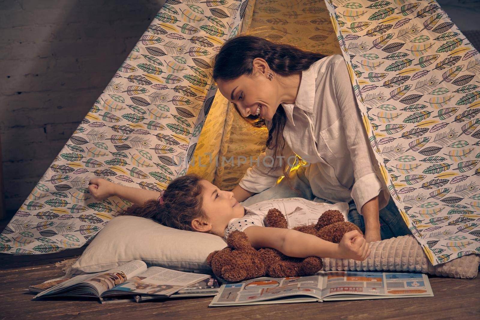 Young mother and her pretty daughter are in a teepee tent with some pillows. Girl is lying with a toy bear. Mom is looking at her and smiling. Happy family.