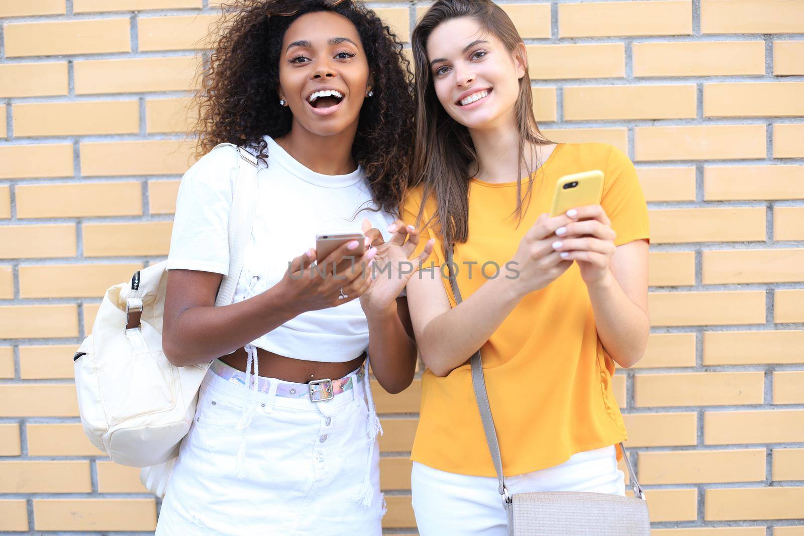 Two young smiling hipster women in summer clothes posing on street.Female showing positive face emotions