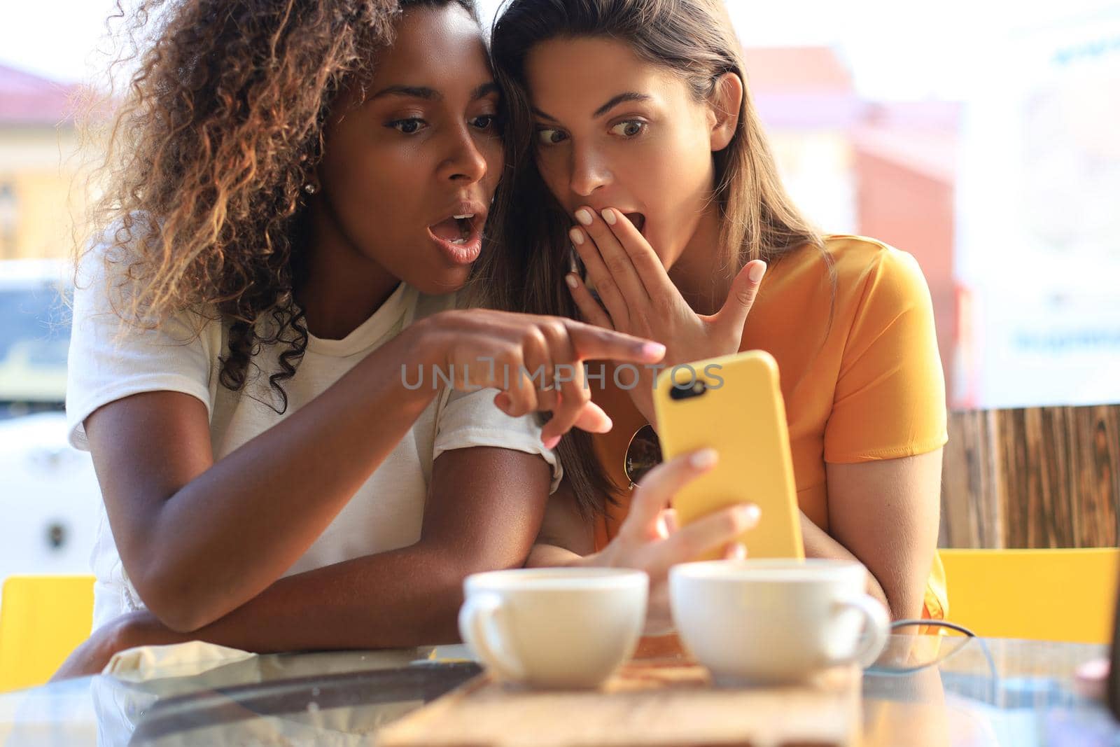 Two beautiful young woman sitting at cafe drinking coffee and looking at mobile phone