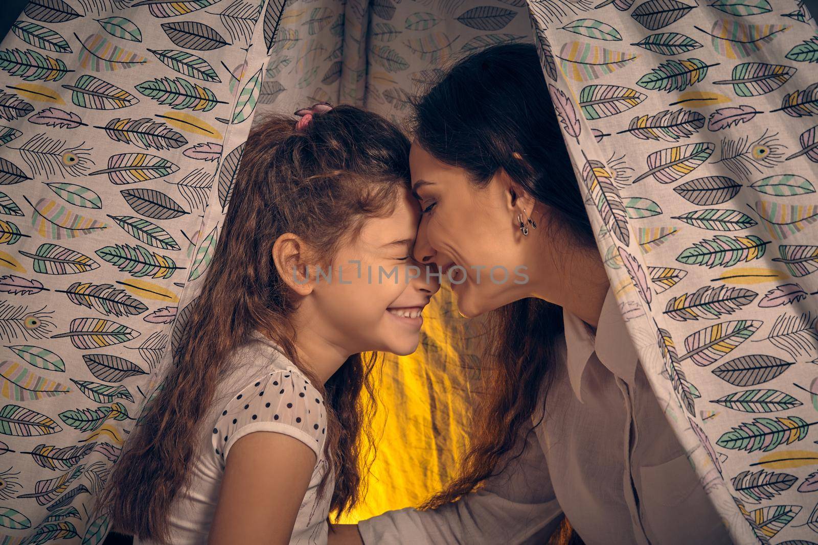 Mother and daughter are sitting in a teepee tent with some pillows, reading stories and laughing. Happy family.