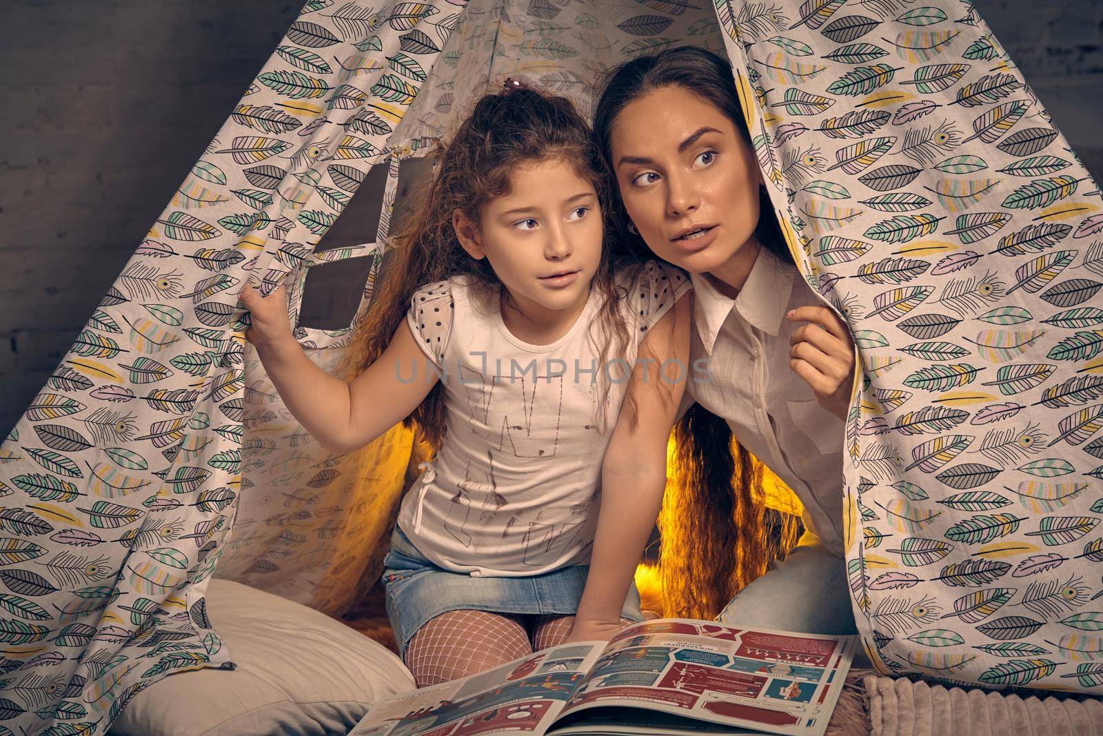 Mother and daughter are sitting in a teepee tent with some pillows and reading stories. Happy family. by nazarovsergey