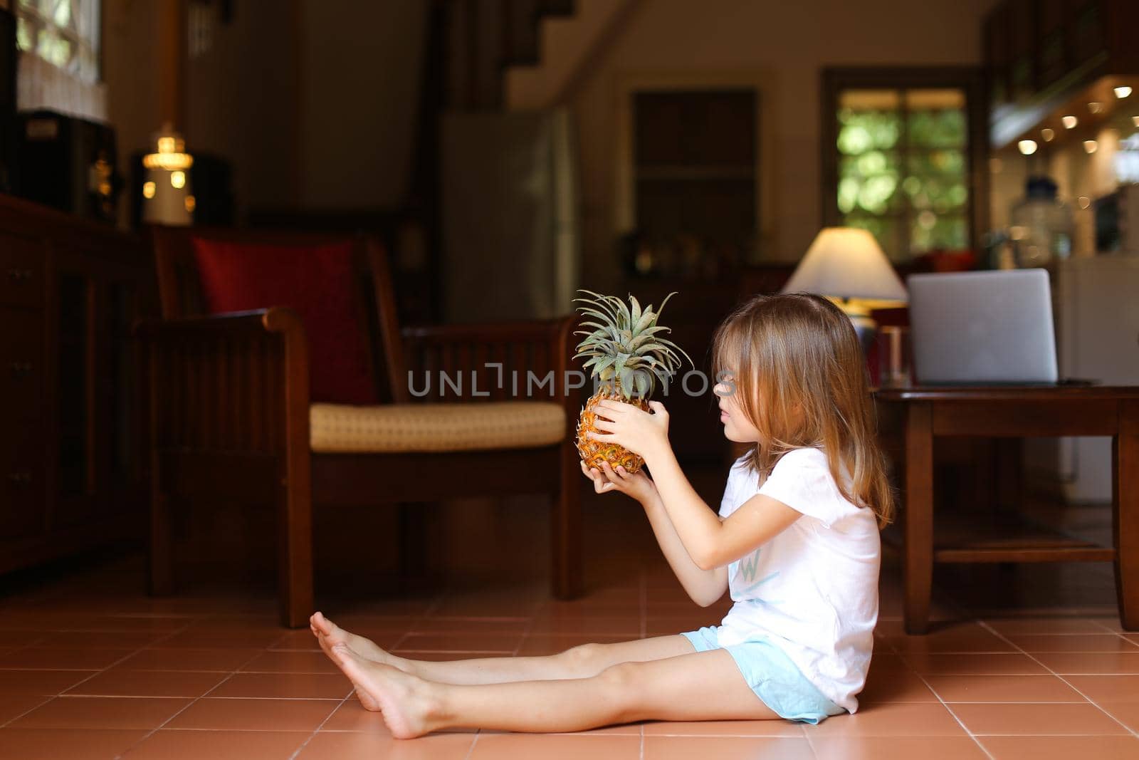 Little female american kid sitting on floor in living room and playing with pineapple, laptop in background. Concept of health life, fruit and childhood.