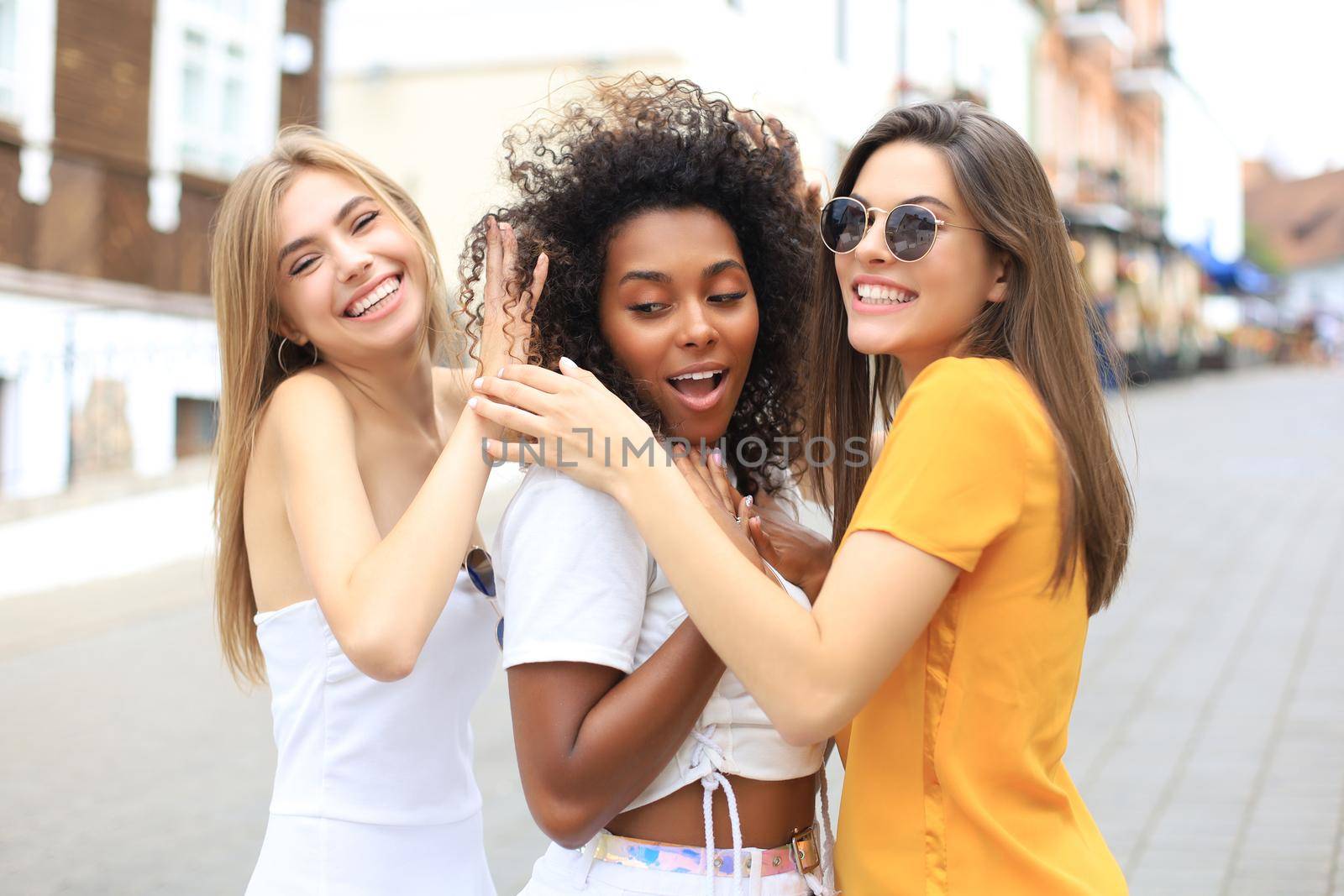 Three young smiling hipster women in summer clothes posing on street.Female showing positive face emotions