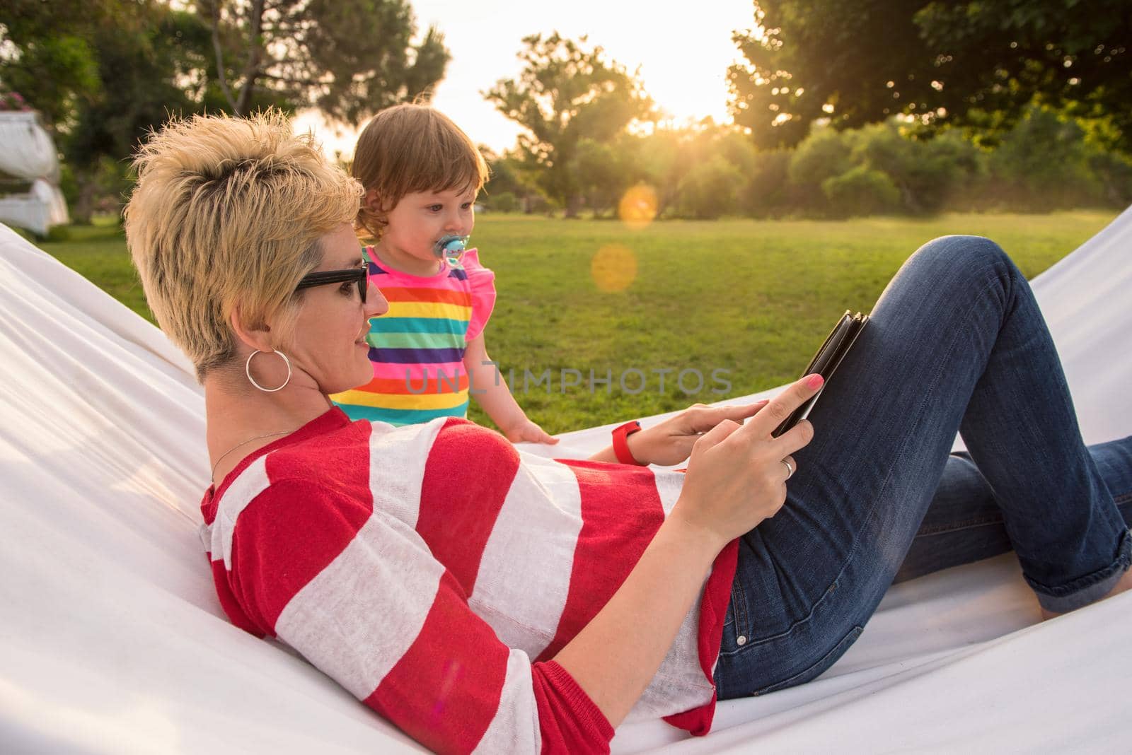 Happy mother and her little daughter enjoying free time using tablet computer while relaxing in a hammock during sunny day on holiday home garden