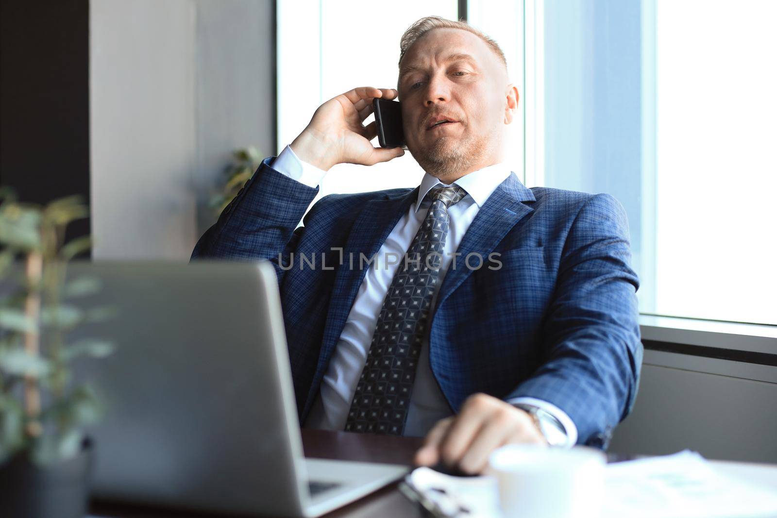 Middle aged businessman with laptop talking on telephone in modern office