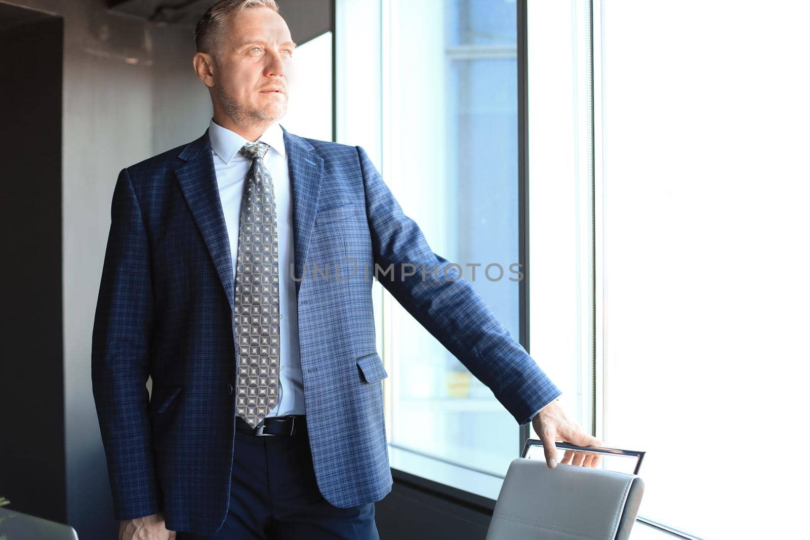 Mature businessman in a corporate suit standing in office and looking away through large windows optimistically