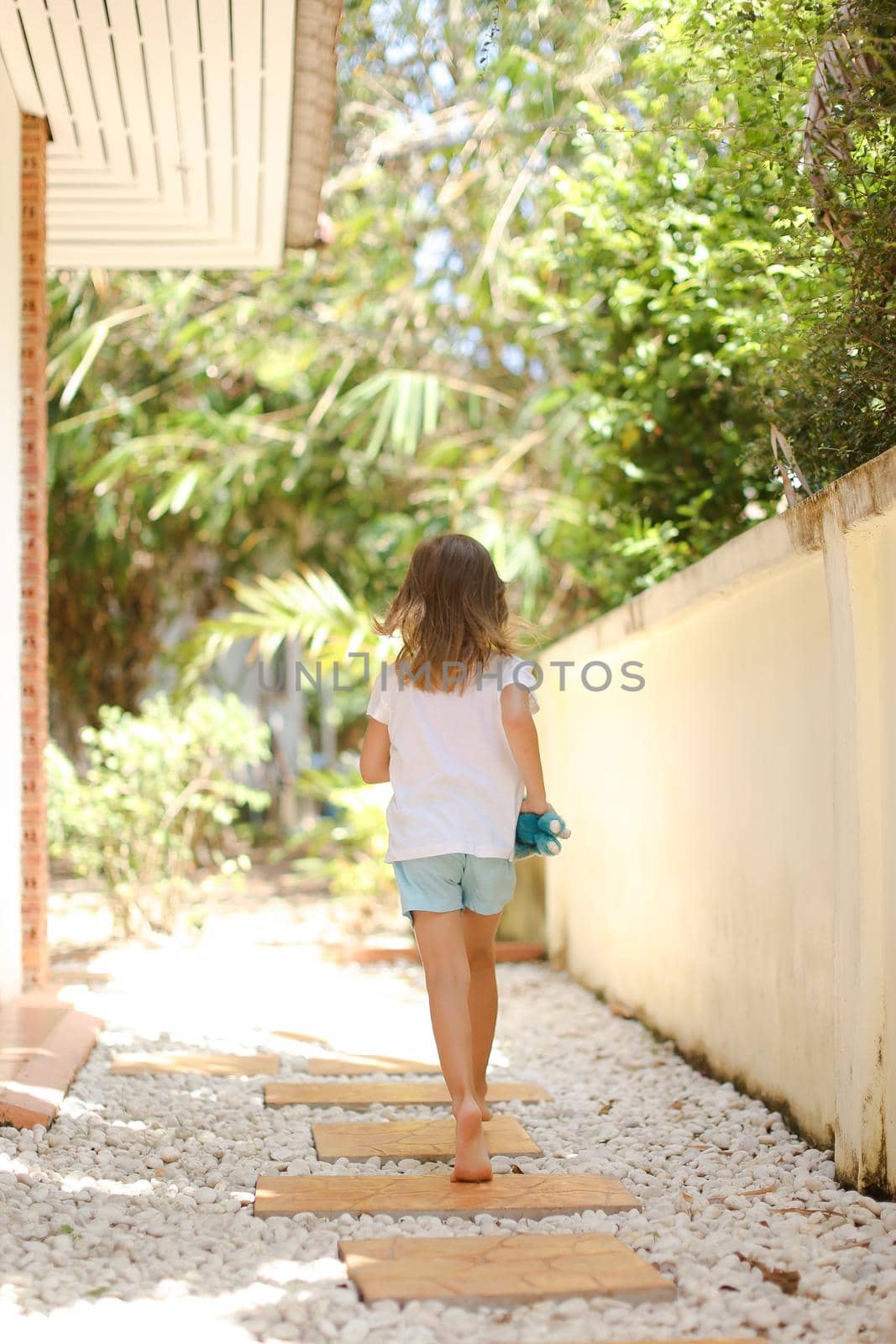 Back view little caucasian female kid walking barefoot on white gravel near exotic hotel. Concept of vacation with kid in Thailand, Phuket.