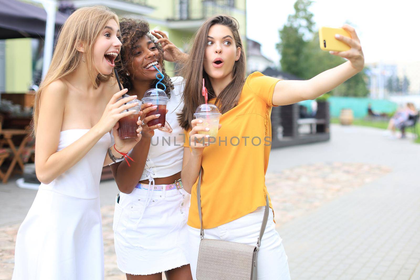 Three trendy cool hipster girls, friends drink cocktail and taking selfie in city background