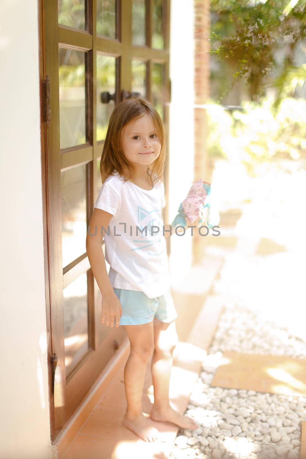 Little female happy kid standing near door with toy outside. Concept of resting with kids in Thailand, exotic hotel.