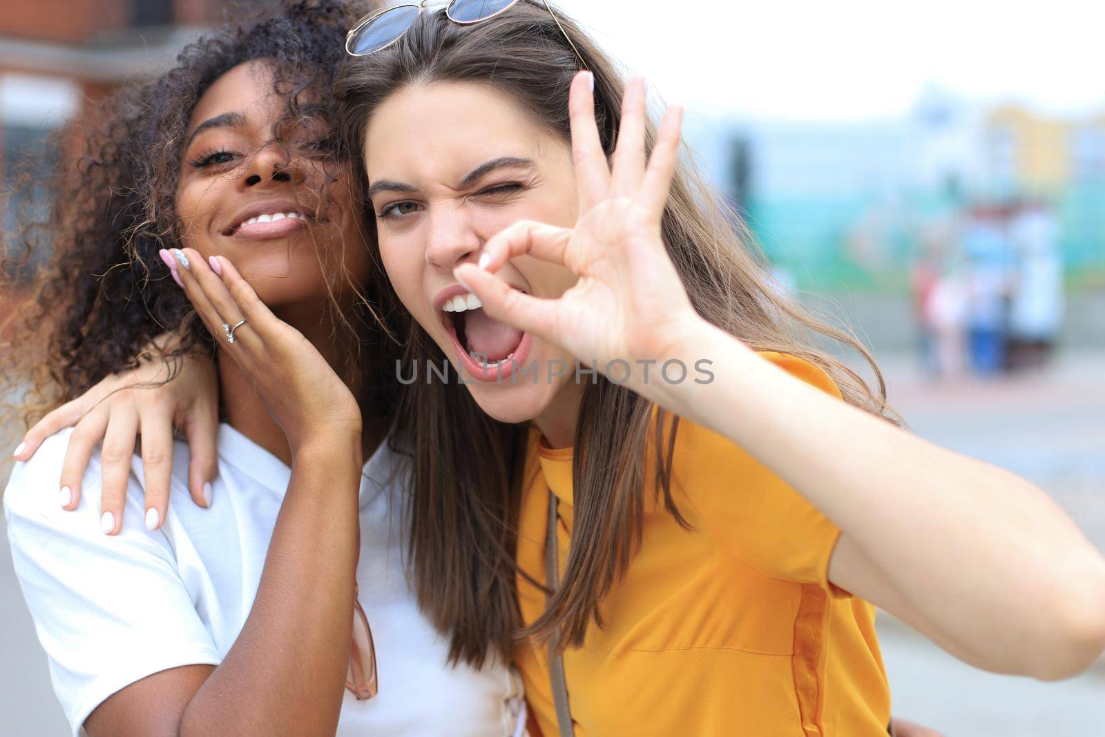 Two young smiling hipster women in summer clothes posing on street.Female showing positive face emotions