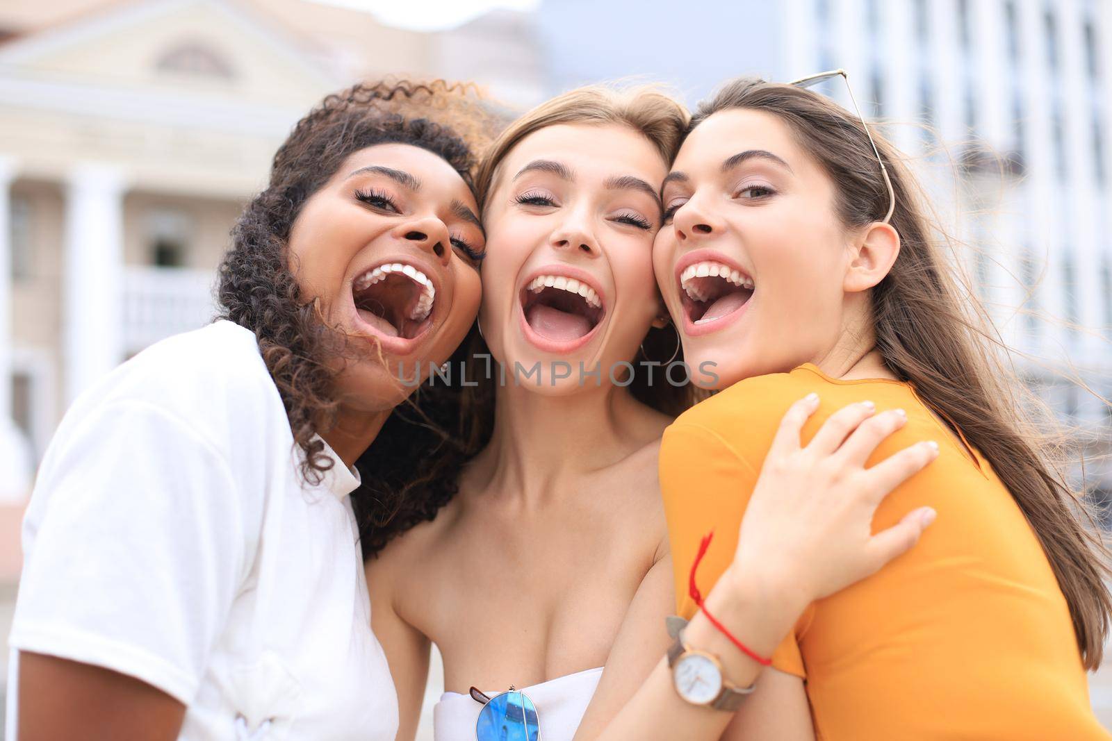 Three young smiling hipster women in summer clothes posing on street.Female showing positive face emotions