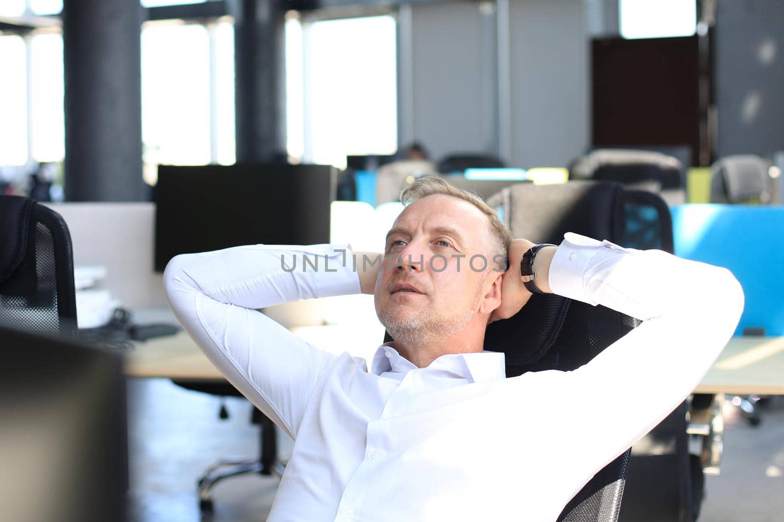 Middle aged modern businessman keeping hands behind head and smiling while sitting in the office. by tsyhun