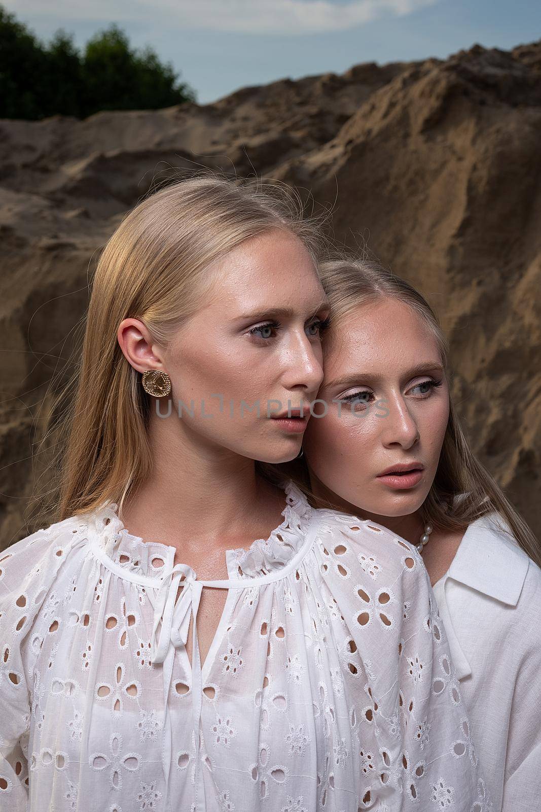 two young blond pretty twins posing at sand quarry in elegant white clothes. stylish glamorous fashion photoshoot with flashlight. girl holds her head on shoulder of her sister in front of sand wall