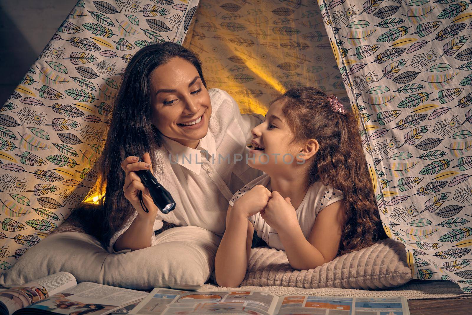 Mother and her pretty daughter are sitting in a teepee tent, reading stories with the flashlight and smiling. Happy family.