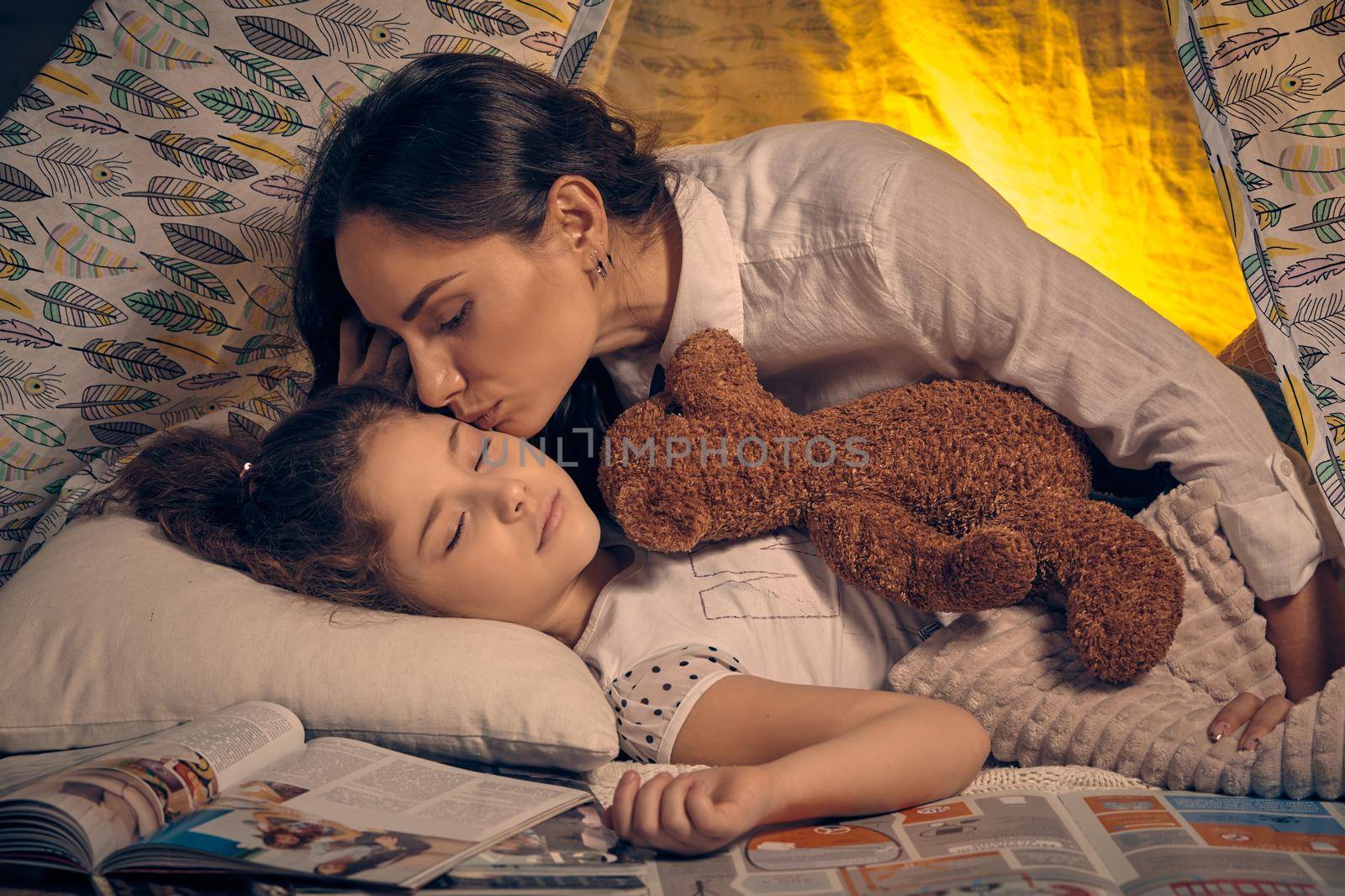 Young mother and her pretty daughter are in a teepee tent with some pillows. Girl is sleeping with a toy bear. Mom is kissing her. Happy family.