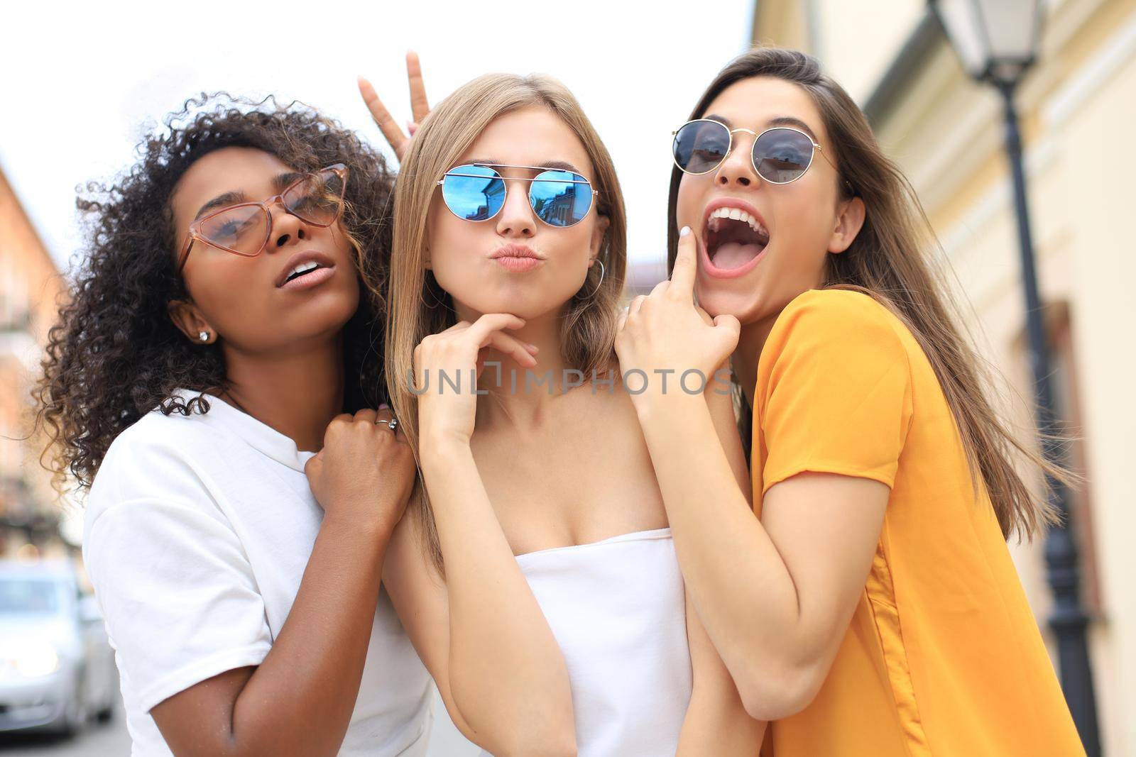 Three young smiling hipster women in summer clothes posing on street.Female showing positive face emotions