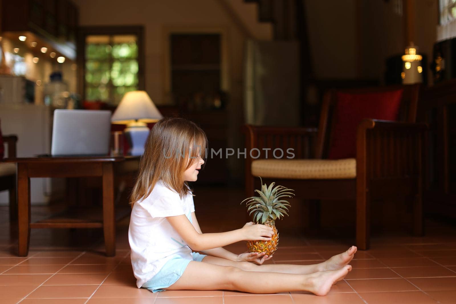Little female kid sitting on floor in living room and playing with pineapple, laptop in background. Concept of health life, fruit and childhood.