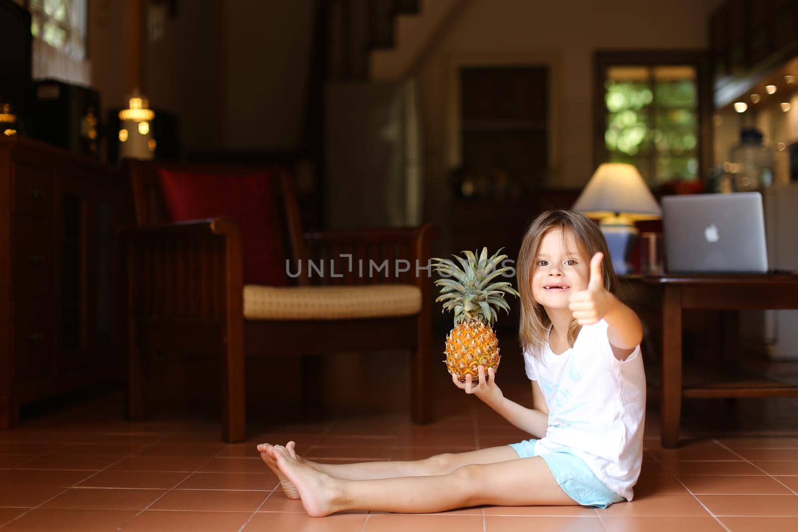 Little smiling female kid sitting on floor in living room with pineapple and showing thumbs up, laptop in background. Concept of health life, fruit and childhood.