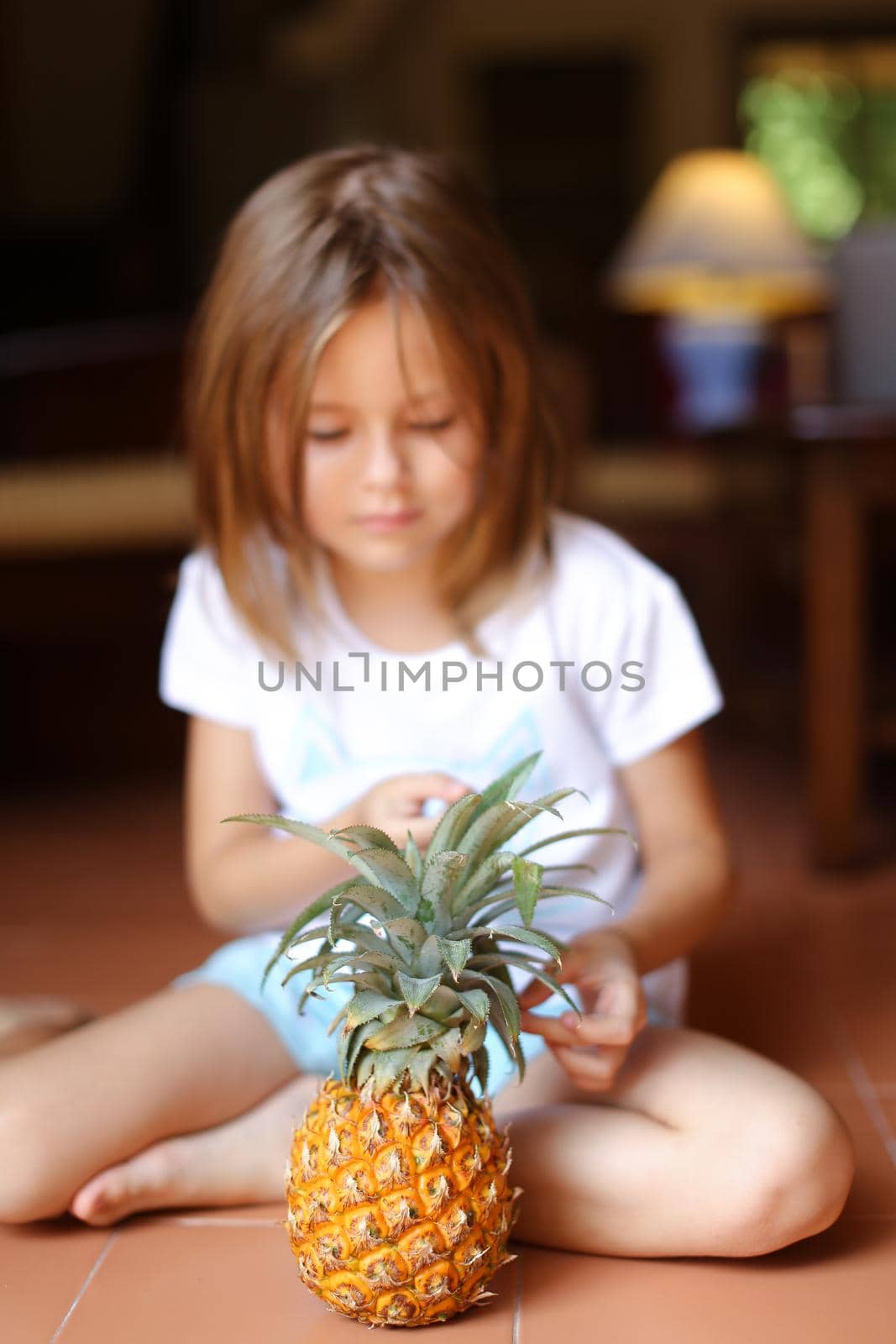Closeup pineapple with little girl sitting barefoot on floor in background. Concept of health life, fruit and kids.