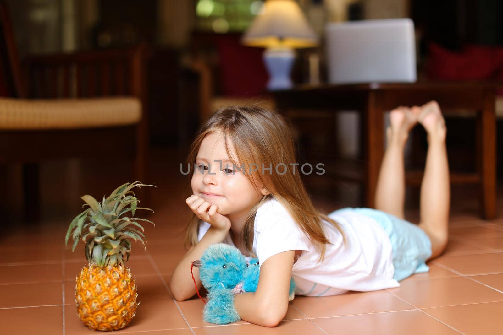 Little barefoot girl lying on floor and playing with pineapple and toy, laptop in background. Concept of health vegeterian life and childhood.