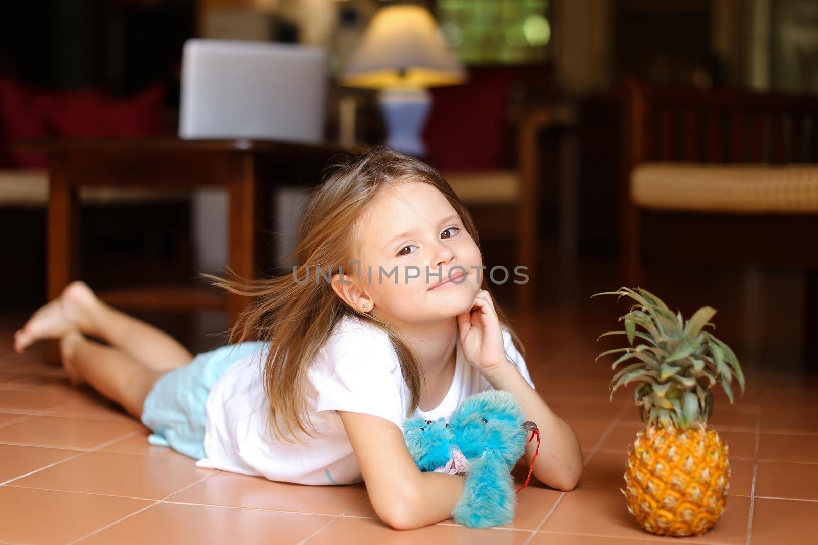 Little barefoot cute girl lying on floor and playing with pineapple and toy, laptop in background. Concept of health vegeterian life and childhood.