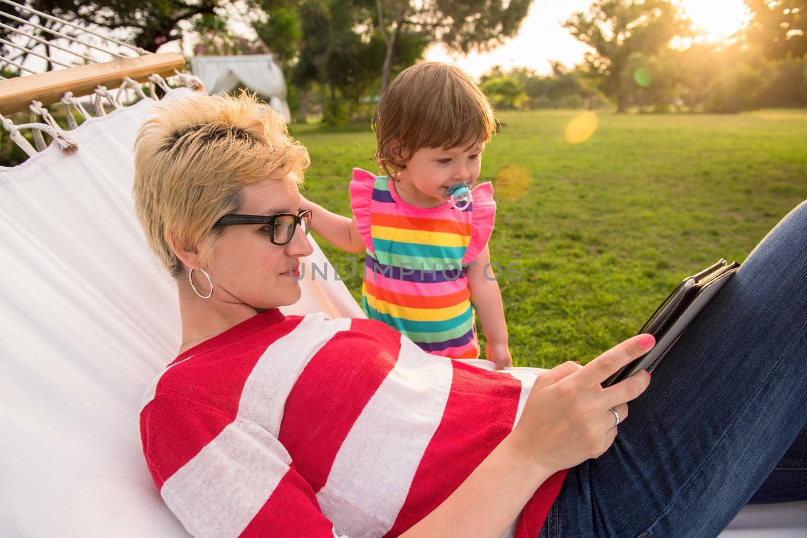 Happy mother and her little daughter enjoying free time using tablet computer while relaxing in a hammock during sunny day on holiday home garden